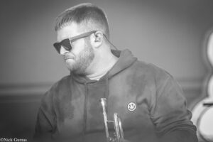Black-and-white photo of a man in a hoodie wearing sunglasses, playing a trumpet on stage, with an in-ear monitor visible in his ear.