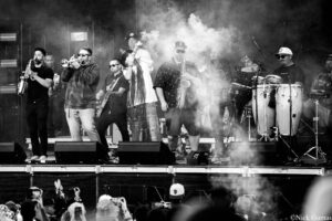 Band of musicians on stage playing brass, woodwinds, and percussion amid stage smoke at a live concert.