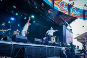 Outdoor concert stage with musicians performing; guitarist left, singer center raising hand, colorful Cal Roots banner above; blue stage lights illuminate the scene