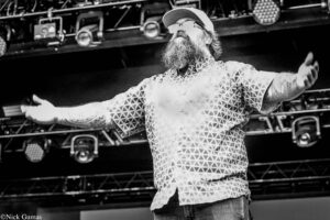 Bearded performer on stage with arms outstretched, wearing a geometric-patterned shirt and cap (black and white).