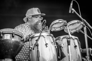Bearded man in a cap and glasses playing a drum set on stage, microphone overhead.