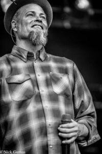 Smiling man with a beard and hat, wearing a plaid shirt, holding a microphone on stage in black and white.