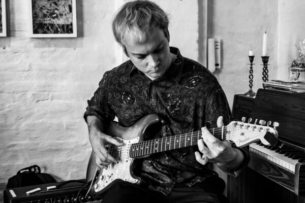 Man in a patterned shirt plays an electric guitar seated in a cozy studio, focusing on the fretboard while an amp sits nearby.