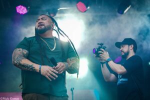 Singer with dreadlocks and tattoos on arms performs on a foggy stage, microphone in hand, while a cameraman films beside him.