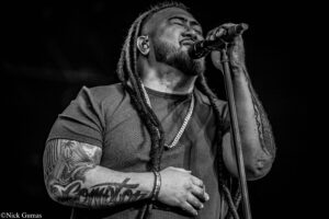 Male singer with dreadlocks singing into a microphone on stage, intense expression, tattoos visible on arms (black-and-white photo).