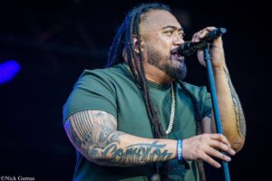 Male singer gripping a microphone on stage, wearing a green shirt and gold chain, with dreadlocks and tattooed arms visible against a dark background.