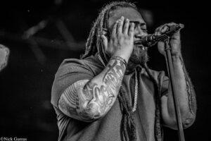 Singer with dreadlocks sings into a microphone, hands pressed to his face, tattoos visible on arms in black-and-white.