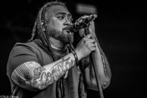 Black-and-white close-up of a tattooed man with dreadlocks singing into a handheld microphone on stage, dark background behind him.