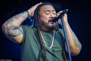Male singer with dreadlocks, eyes closed, hand on his head, singing into a microphone on stage with tattooed arms visible.