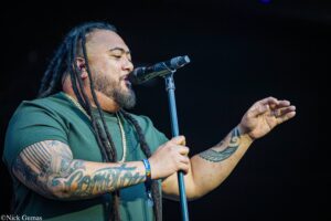 Male performer singing into a microphone on stage, shown in profile with tattoos on his arms and dreadlocks visible.