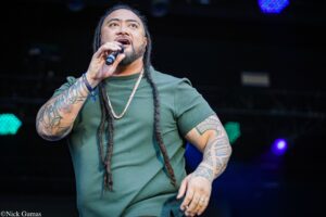 Male singer with braided hair and full arm tattoos performing on stage, microphone in hand.