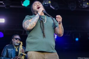 Male singer with long dreadlocks and tattoos belts out into a handheld microphone on stage during a concert, wearing a green shirt and gold chain.