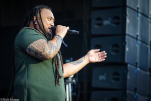Man with dreadlocks singing into a microphone on stage, wearing a green shirt and visible tattooed arms.