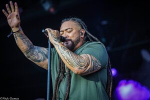 Singer with tattoos and dreadlocks passionately performs on stage, hand raised, gripping a microphone close to his mouth.