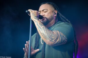 Male singer with dreadlocks and full-arm tattoos singing into a microphone on stage.