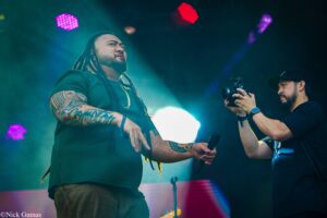 Live concert scene with a tattooed singer in a green shirt and gold chain, facing the audience as a cameraman films from the right under colorful lights.