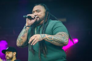 Male performer with dreadlocks wearing a green shirt singing into a microphone on stage, tattooed arms visible against purple lighting.