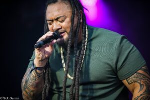Singer with dreadlocks and tattoos singing into a microphone on stage with purple lights in the background, eyes closed in concentration.