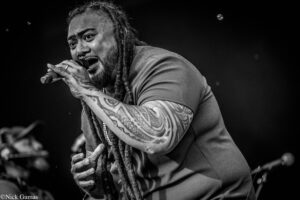 Performer with dreadlocks and tattooed arms singing into a microphone on stage (black and white).