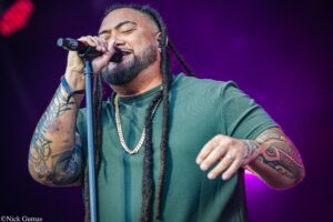 Male singer with dreadlocks performs into microphone on stage, purple backdrop and spotlight in view; wearing a green shirt and gold chain.