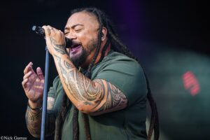 Male singer with braided hair and tattooed arms performing into a microphone on stage.