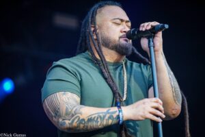 Male singer with dreadlocks wearing a green shirt, singing into a microphone on stage.
