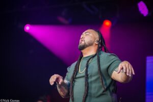 Male performer with long dreadlocks on stage under purple lighting, eyes closed, mid-sentence singing into microphone (live concert).