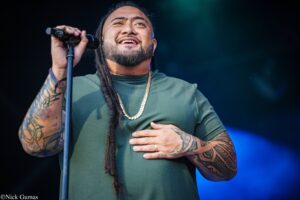 Male singer with braided hair and a beard, wearing a green shirt and gold chain, singing into a microphone on stage, tattoos visible on arms.