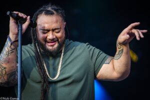 Male singer with dreadlocks and tattoos, holding a microphone and singing with eyes closed on stage.
