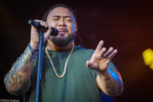 Male singer with closed eyes, gripping a microphone on stage, wearing a green shirt and gold chain.