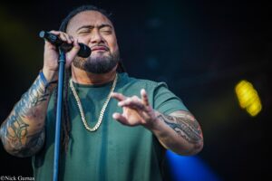 Male singer with tattoos and a gold chain passionately singing into a microphone on stage, wearing a green shirt.