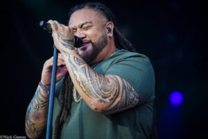 Tattooed male singer singing into a microphone on stage, wearing a green shirt and a gold chain.