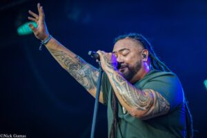 Male singer with braided hair and full-arm tattoos raises his left hand while singing into a microphone on stage.