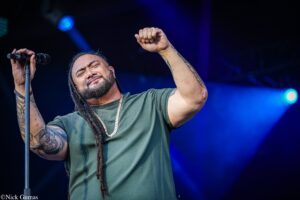 Male singer on stage raising a clenched fist while holding a microphone, wearing a green shirt and chain, with blue stage lights in the background