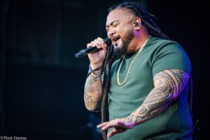 Male singer with dreadlocks passionately singing into a handheld microphone on stage, wearing a green shirt and a gold chain.