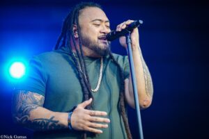Male singer with dreadlocks singing into a microphone on stage, wearing a green shirt and gold chain, blue concert lighting behind him.