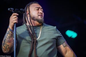 Male singer with long dreadlocks on stage, gripping a microphone stand, wearing a green shirt and gold chains.