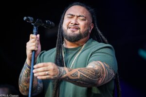 Male singer with eyes closed, gripping a microphone on stage, wearing a green shirt and gold chain, with long dreadlocks and tattooed arms.
