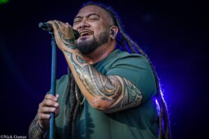 Male vocalist with tattoos and dreadlocks singing into a microphone on stage, purple lights behind him.