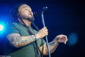 Male singer with dreadlocks and tattoos performing into a microphone on stage with blue lighting in the background.
