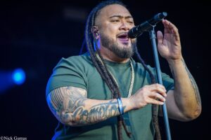 Male singer with dreadlocks and tattoos singing into a microphone on stage, wearing a green shirt and gold chain.