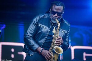 Jazz saxophonist in sunglasses and a black leather jacket performs on a blue-lit stage, saxophone in hand.
