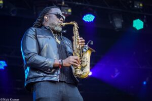 Saxophonist wearing sunglasses and a dark jacket performs on a dim stage with blue stage lights in the background