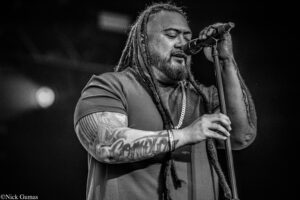 Male singer with dreadlocks and tattoos singing into a microphone on stage, eyes closed, in black and white.