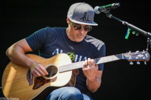 Man wearing sunglasses and a gray cap with 'hi' logo plays an acoustic guitar on stage, microphone overhead.
