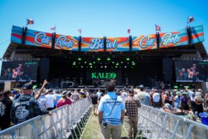 Outdoor music festival stage with bright Cali Roots banners and a crowd behind metal barriers, sunny day