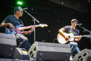 Two male musicians perform on a dark stage: one bass guitarist on the left and a singer with an acoustic guitar on the right, with bubbles in the air.