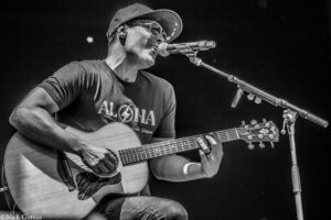 Male guitarist and vocalist wearing a cap and sunglasses, performing on stage with an acoustic guitar and microphone (black‑and‑white photo).