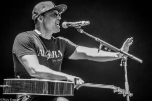 Male musician wearing sunglasses and a cap, singing into a microphone while playing a cajón on stage, in black and white.