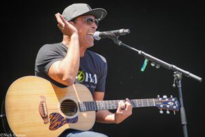 Male musician wearing sunglasses and a cap, playing an acoustic guitar and singing into a microphone on stage.
